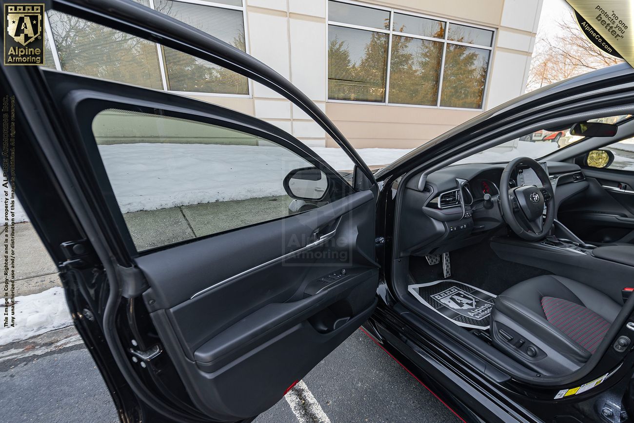 An open driver's side door of a black Toyota Camry by Alpine Armoring  reveals the interior, including the driver's seat, dashboard, and carpeting with protective paper. Frosted lawn and building exterior visible in the background.
