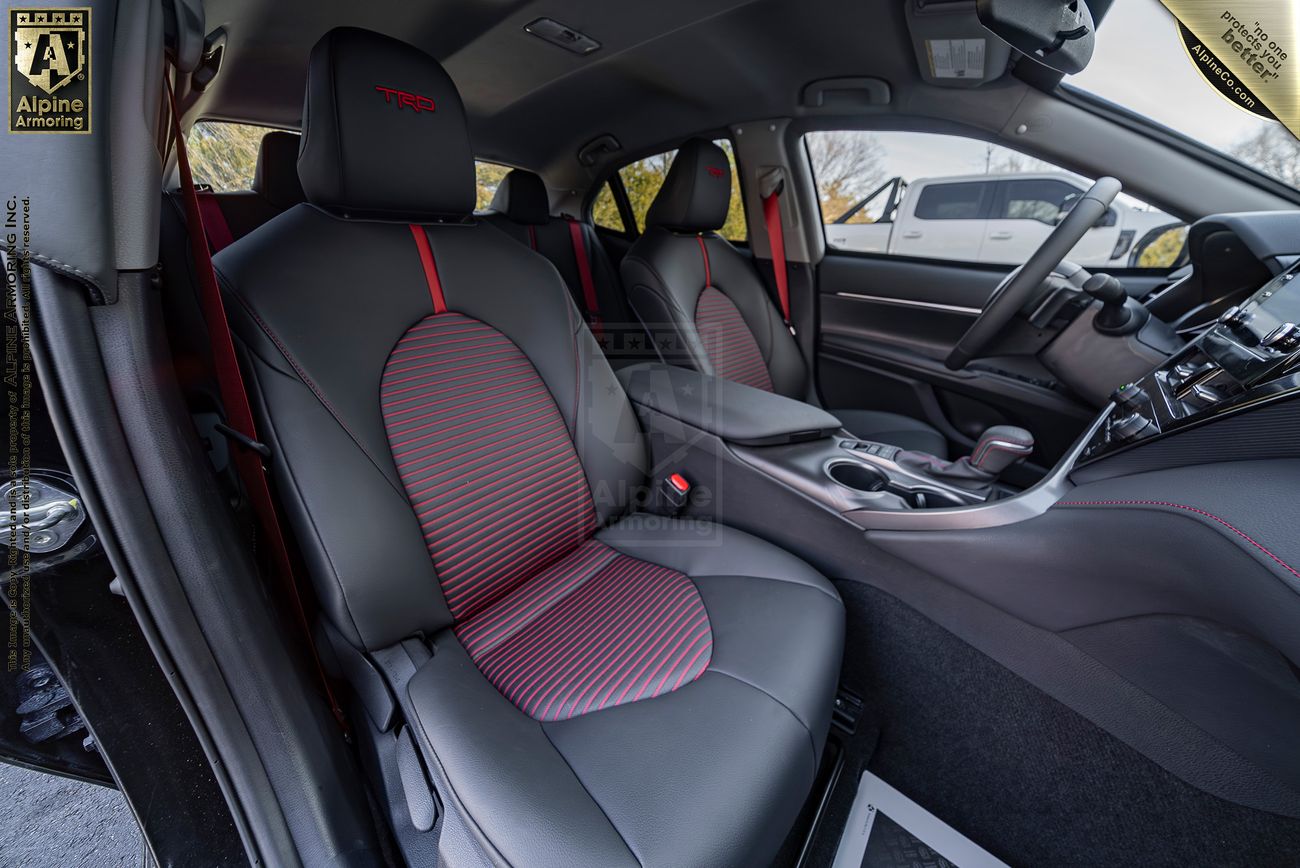 Interior view of a black armored Toyota Camry TRD sedan featuring black and red leather seats, a sleek dashboard with a gear shift, and advanced touchscreen controls. The car door is open, showing part of the exterior.