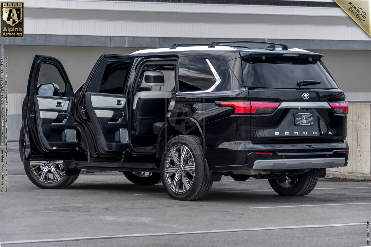 A black armored Toyota Sequoia Capstone from Alpine Armoring, shown with all doors open to reveal the luxurious interior. The vehicle is parked in an outdoor setting with a concrete wall and building in the background.