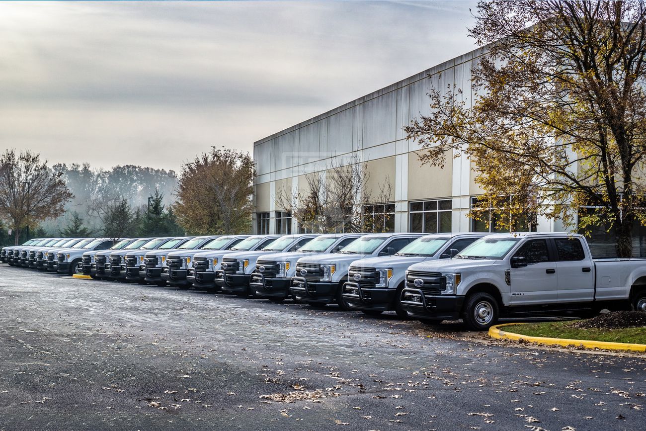 A line of white pickup trucks is parked next to the Alpine Armoring headquarters on an overcast day.
