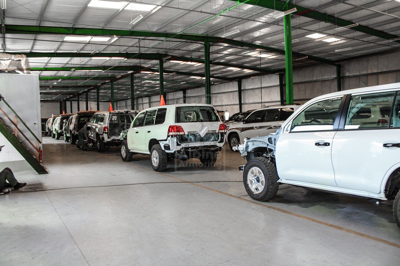 Several partially assembled vehicles are lined up in a large industrial warehouse with green support beams.