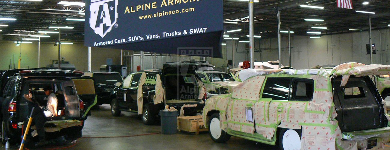 Alpine Armoring facility showing vehicles in various stages of armoring process, including SUVs and vans with partially installed armor plating.