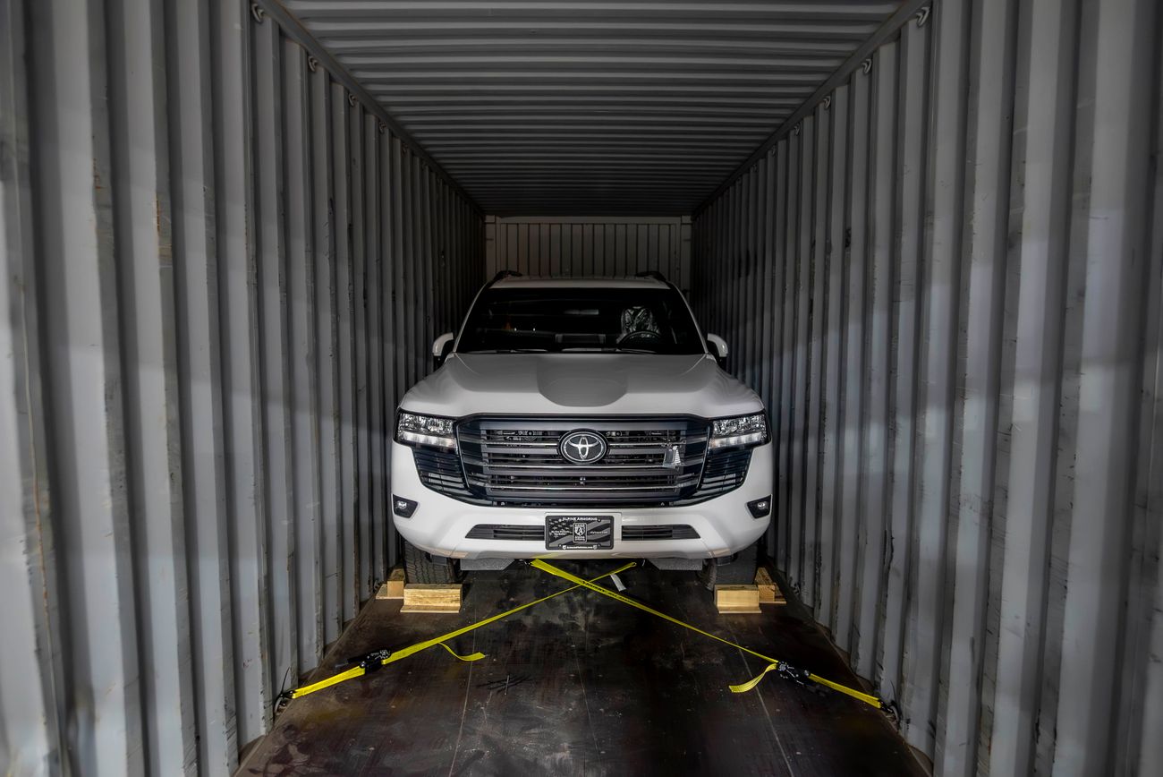 A white SUV secured inside a shipping container with yellow straps and wooden blocks, viewed from the front.