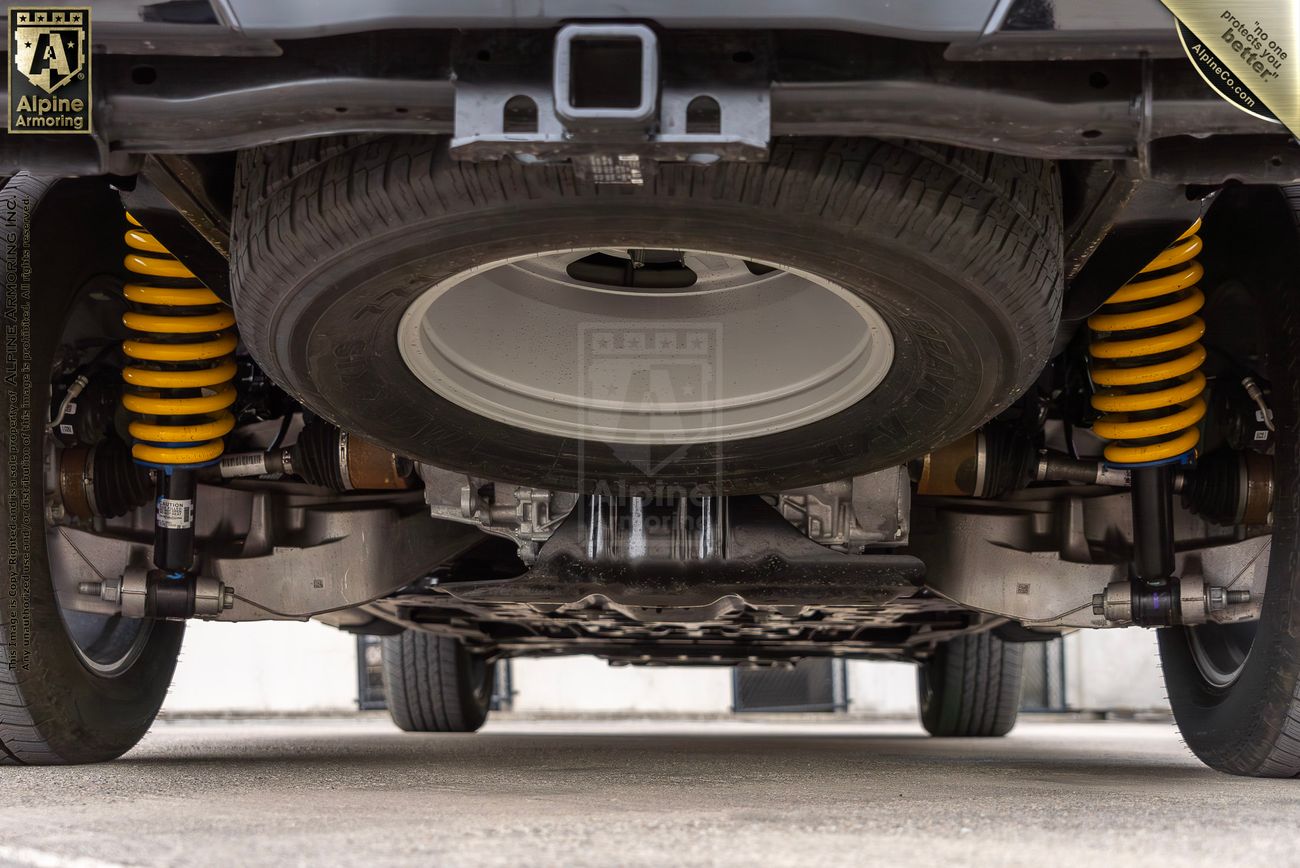 The underside of an armored vehicle from Alpine Armoring, featuring a spare tire, yellow suspension coils, and robust undercarriage components.