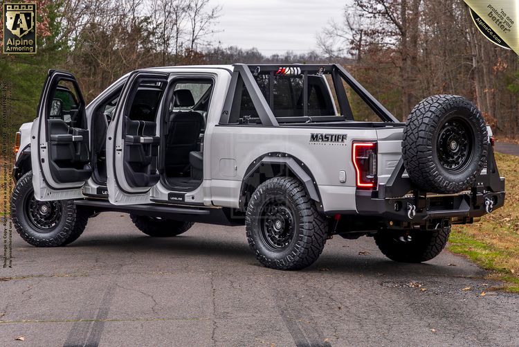 A silver customized Mastiff with off-road tires, open doors, and a spare tire mounted on the back, parked on a paved surface with trees in the background.