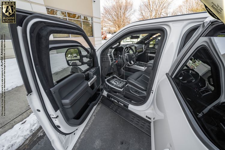 An open driver door shows the interior of a white Mastiff parked outdoors near a snowy sidewalk, with visible trees and a building exterior in the background.