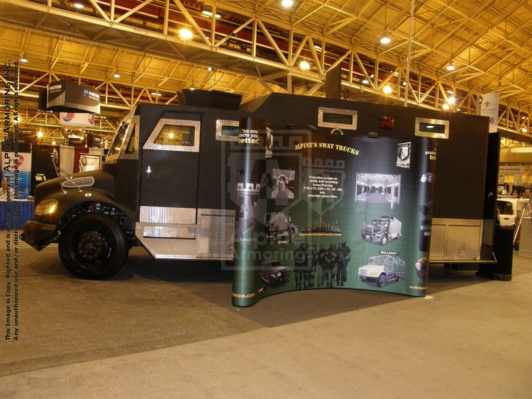 A black armored Alpine Armoring Bulldog displayed indoors at an exhibition, showcasing images and features of SWAT trucks on a poster board attached to its side.