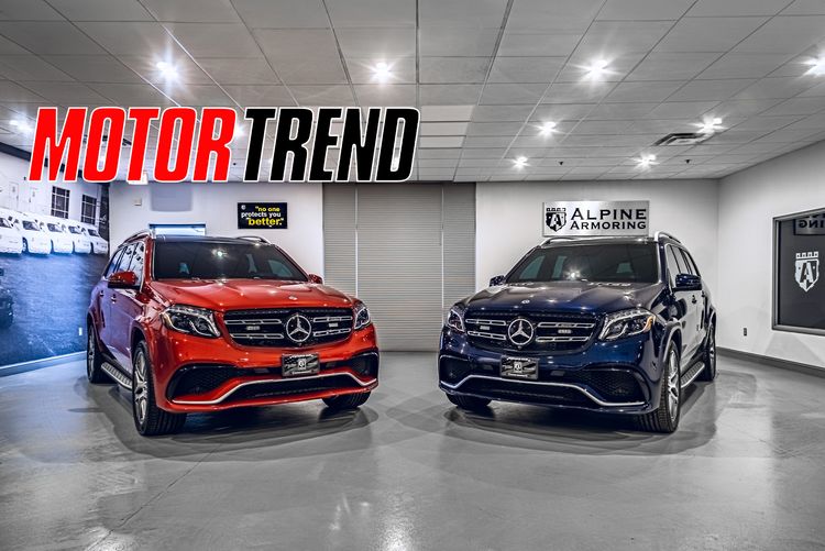 Two armored Mercedes-Benz GLS SUVs in Alpine Armoring showroom, one red and one black, displayed under Motor Trend signage.