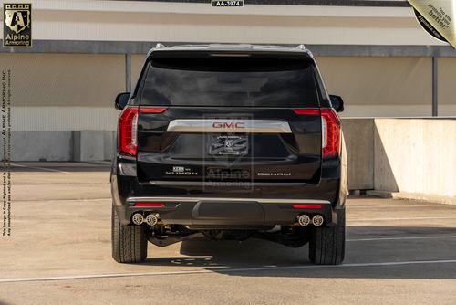 Rear view of a black arnmored GMC Yukon Denali SUV with dual exhaust pipes, displayed in a parking area. The vehicle has "Alpine Armoring" branding on the back.