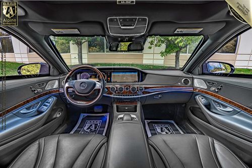 Interior view of armored black Mercedes S550 luxury sedan  featuring a leather-wrapped steering wheel, digital dashboard, wood trim accents, and two front seats separated by a central console. Trees visible through the windshield.